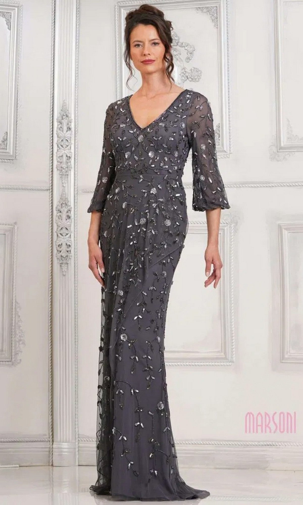 A woman stands before a white ornate wall in the Marsoni MV1320 long formal dress in charcoal, featuring three-quarter sleeves and silver floral embellishments.